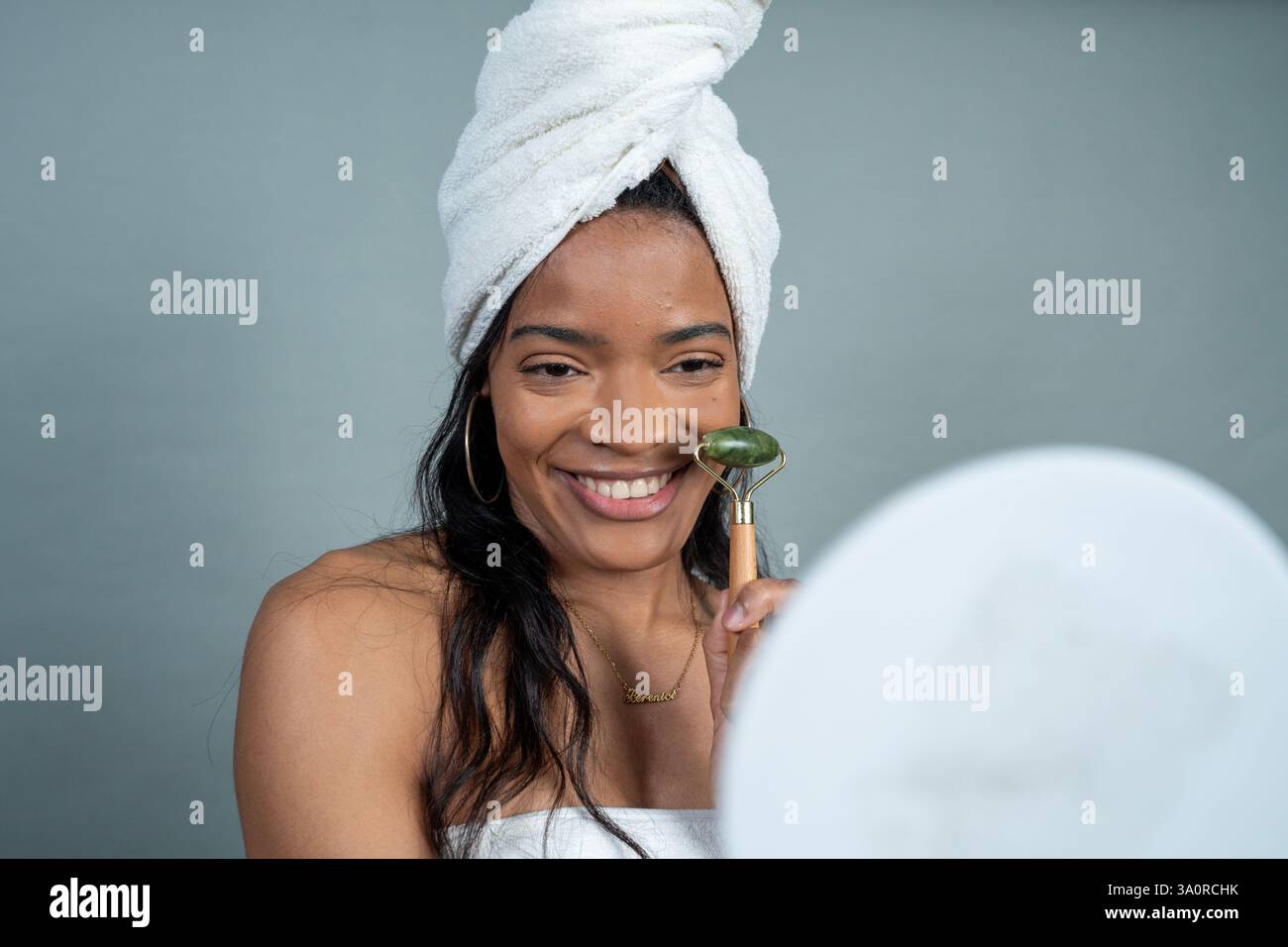 Happy woman wearing towel using jade roller for face massage in front of mirror after shower Stock Photo