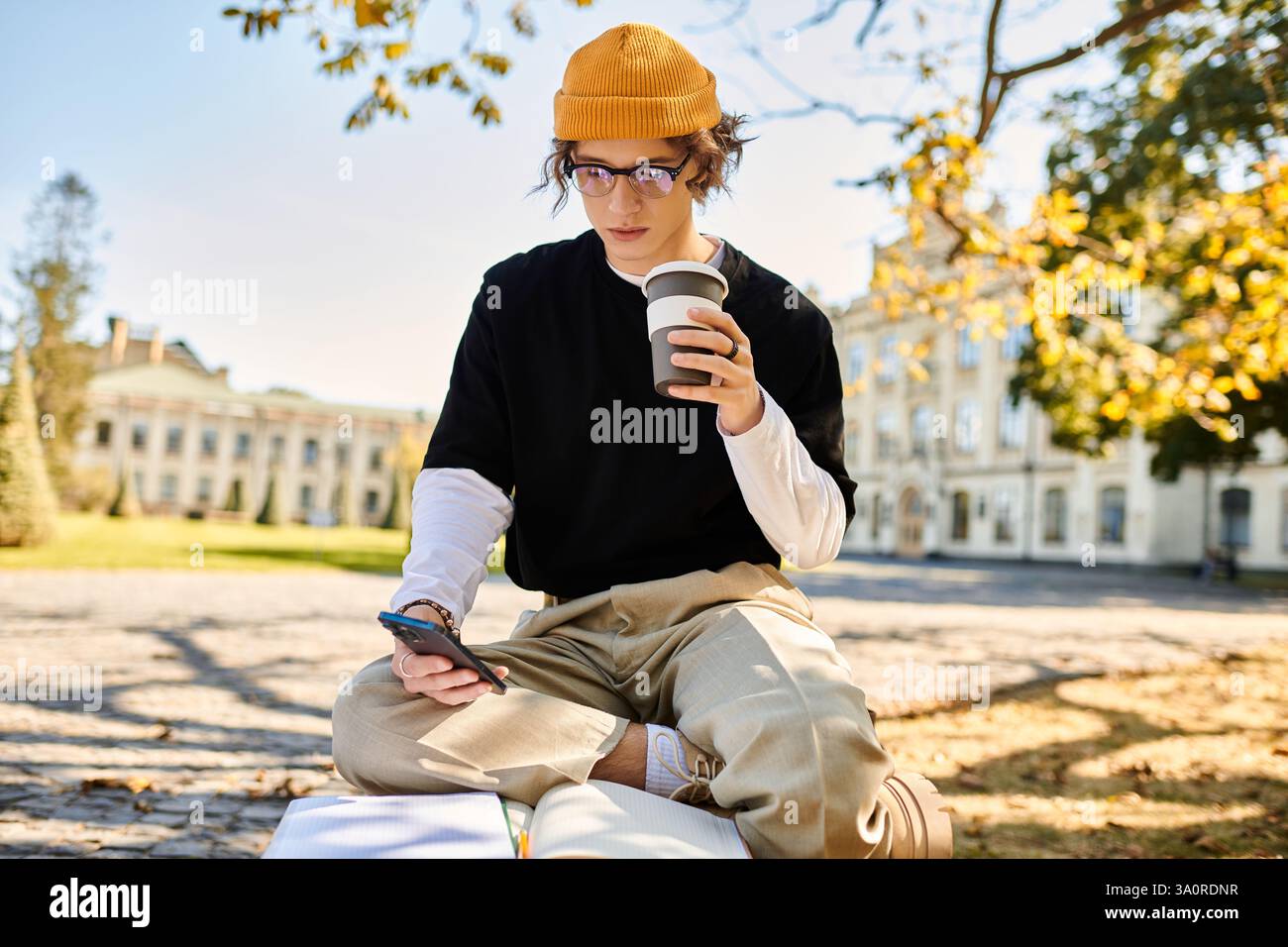Stylish young man sips coffee and checks his phone while sitting on a sunny park bench Stock Photo