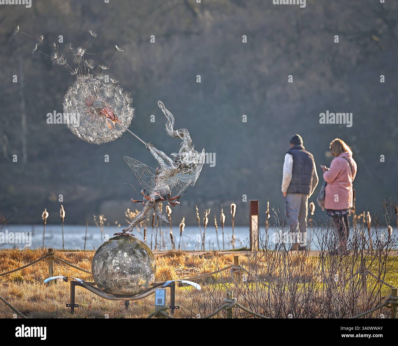 A couple look over the lake at Trentham Gardens in early spring with wire fairy and dandelion sculpture by Robin Wight in the foreground Stock Photo