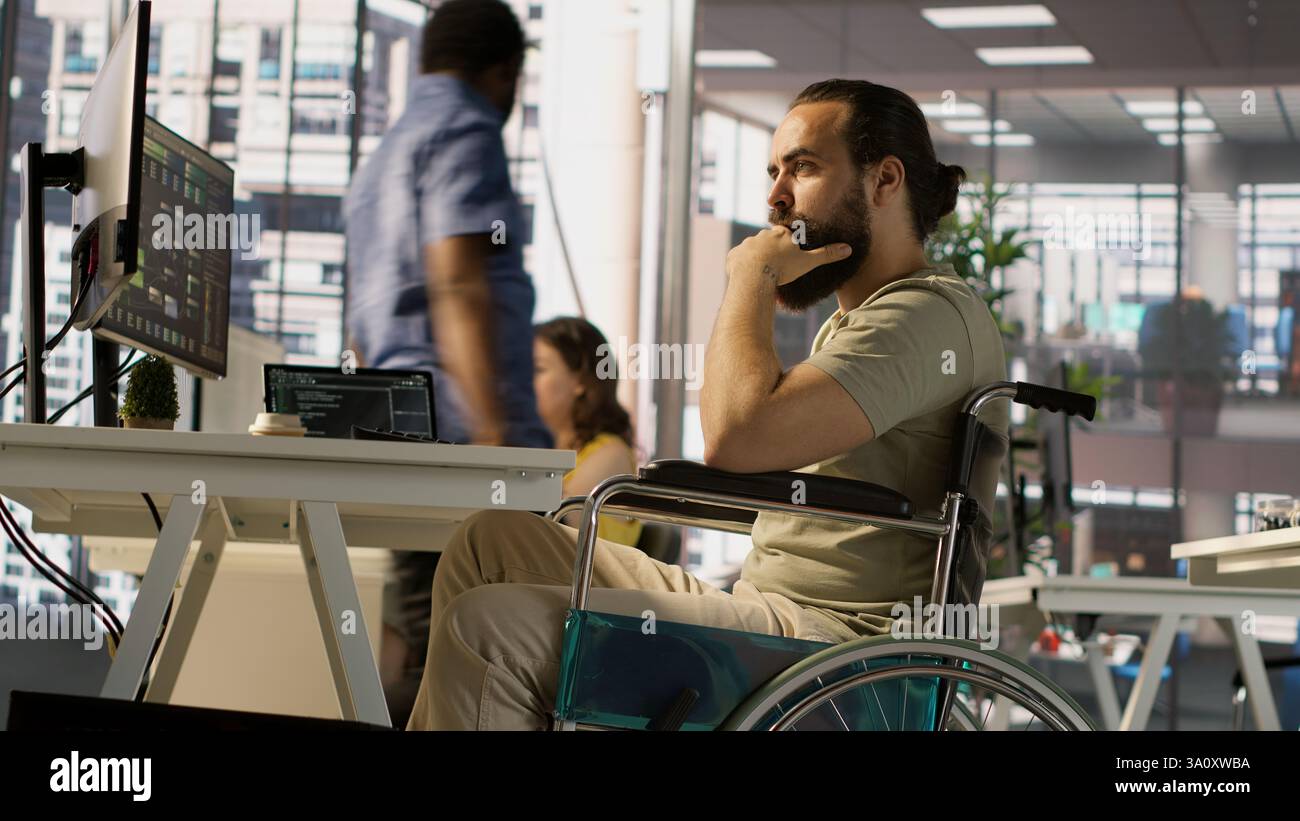 IT worker with disability sitting in wheelchair in office working at computer desk, focused on testing programs. Software engineer with paraplegia in inclusive workplace checking code on PC, camera A Stock Photo