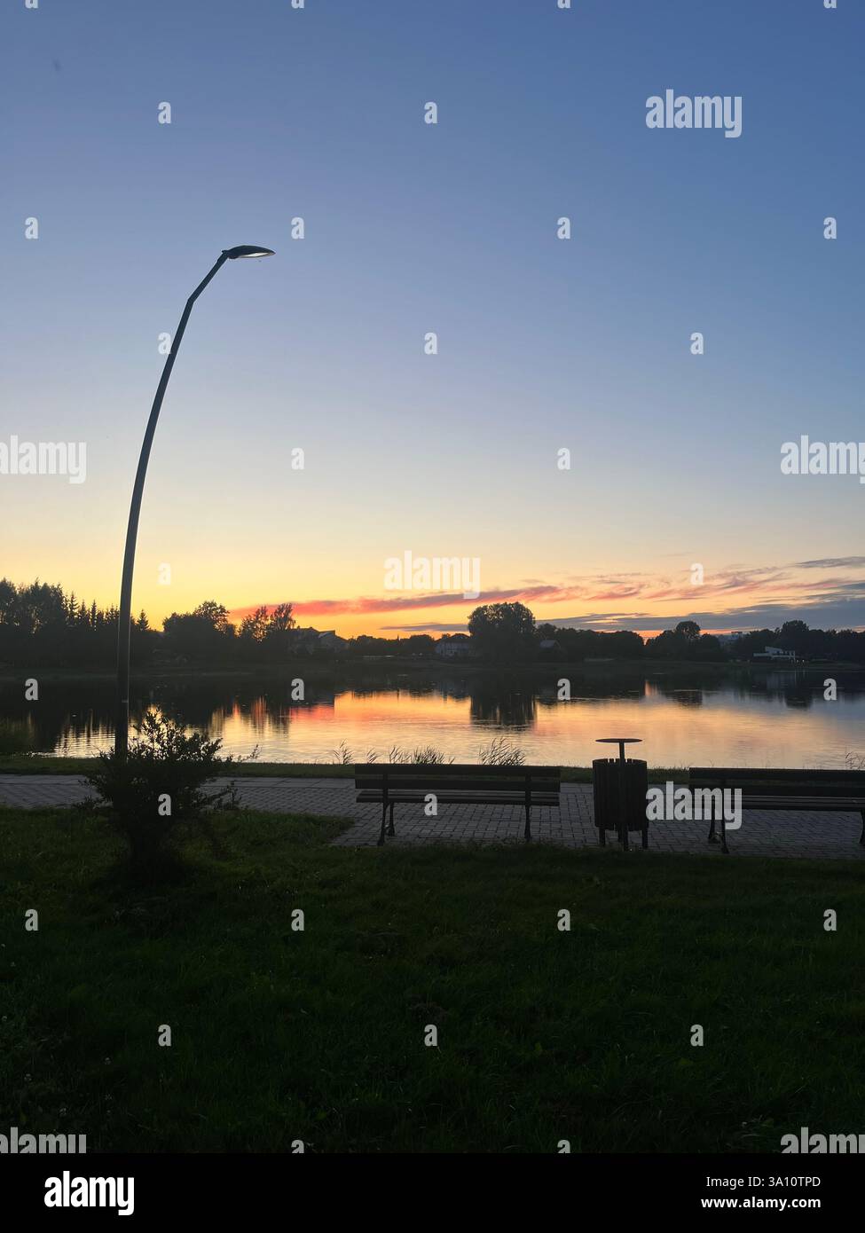 A peaceful evening as the sun sets, casting a warm glow across the scene. In the foreground, a calm lake stretches out, with its shoreline. - Smartphone Captured Stock Image