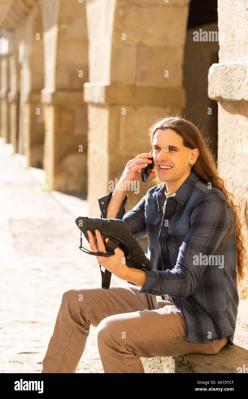 Smiling man talking on smartphone while using a rugged tablet outdoors. Lifestyle, technology. Stock Photo
