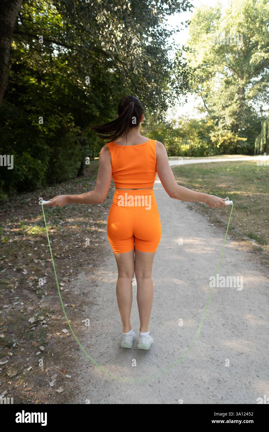 A young woman in a bright orange outfit jumps rope in a green park on a summer morning, staying active and healthy outdoors. Stock Photo
