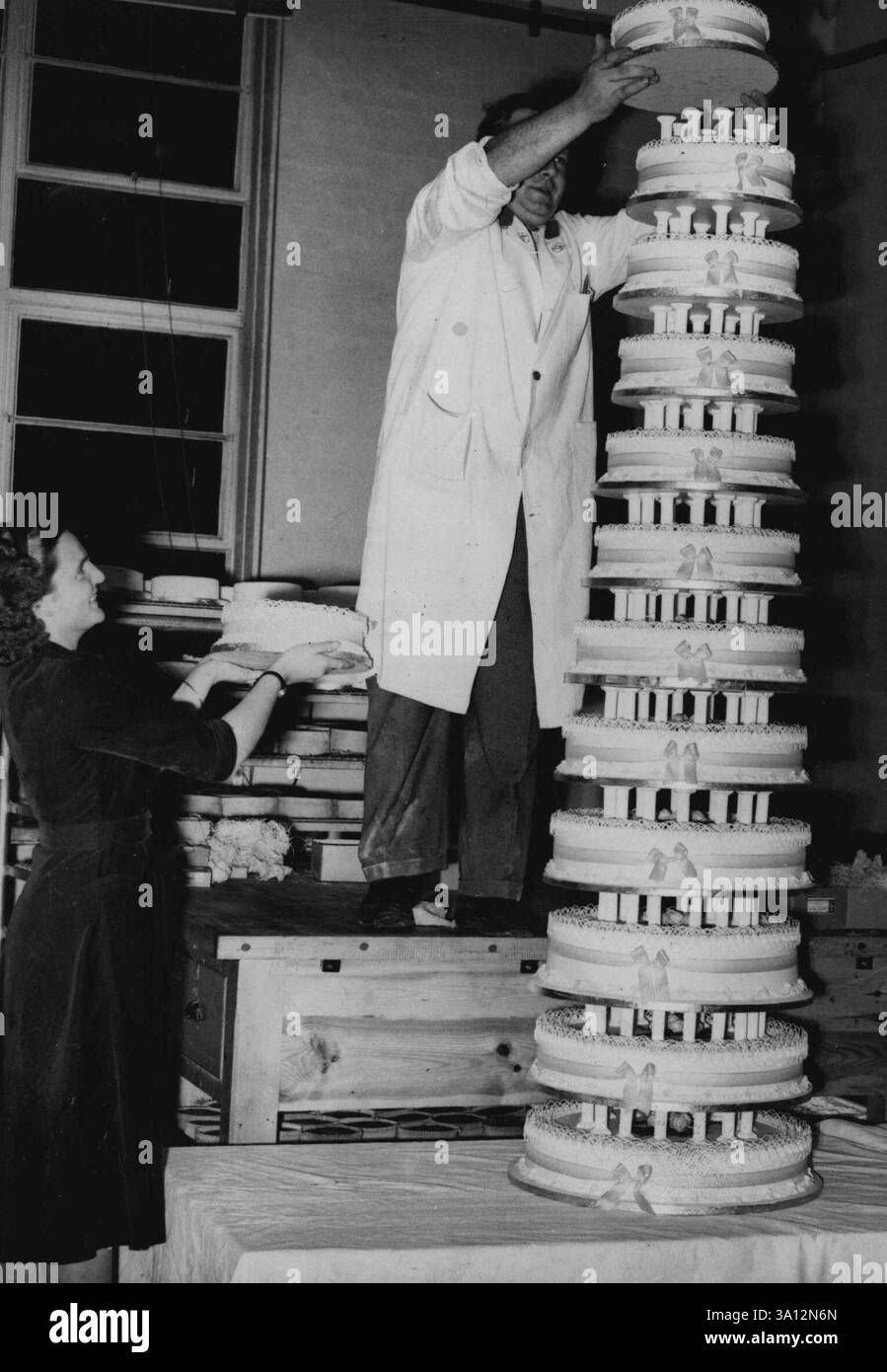 12 Tiers And Still Rising - Barbara Truscott assists in 'building-up' the giant birthday cake. The ceiling was not high enough to take a picture of the cake at its full height.When Barbara Truscott has her 21st birthday party on Monday at Plymouth she will be presented with what may well be the biggest cake in the world. It will be 18 feet high and of 21 tiers.Birthday cake for Barbara Truscott's 21st party at Plymouth, England, was 18ft high, mode in 21 tiers. It was too high to be made in one piece in bakehouse. October 31, 1952. (Photo by Paul Popper, Paul Popper Ltd.). Stock Photo