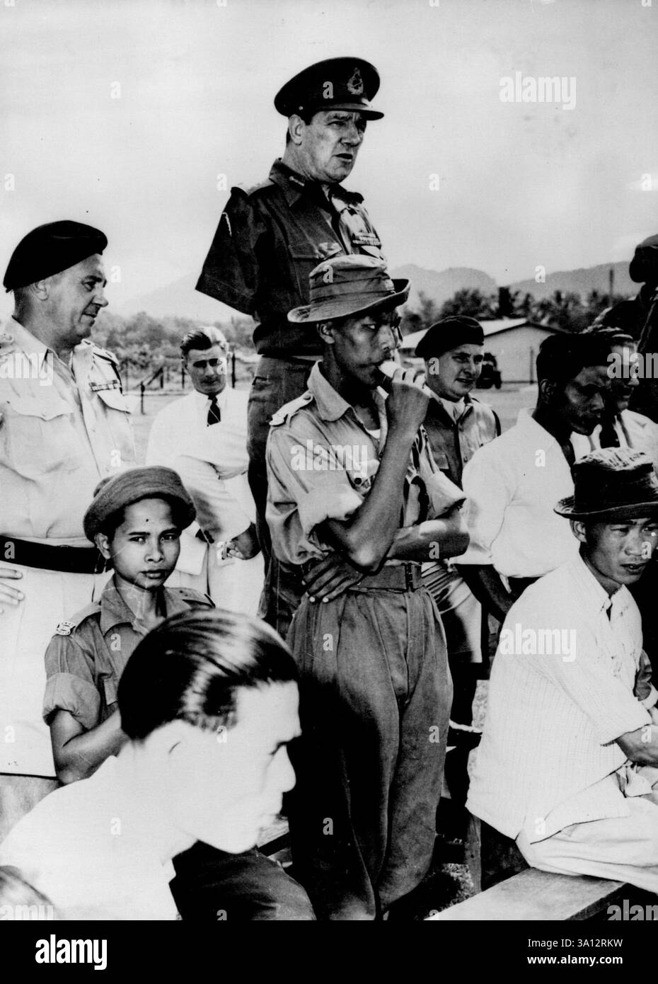 Lollipop Soldier Missed the General:This Malayan home guard was so intent on his iced lollipop and the shooting at the Perak state home guard shooting championships recently (early July) that he did not notice his two important fellow spectators. The one standing on a seat for a better view is Lieutenant General G. K. Bourne, New Director of operations, Malaya. At left is Brigadier I.A.C. Lauder, state home guard officer. July 20, 1954. (Photo by Associated Press Photo). Stock Photo