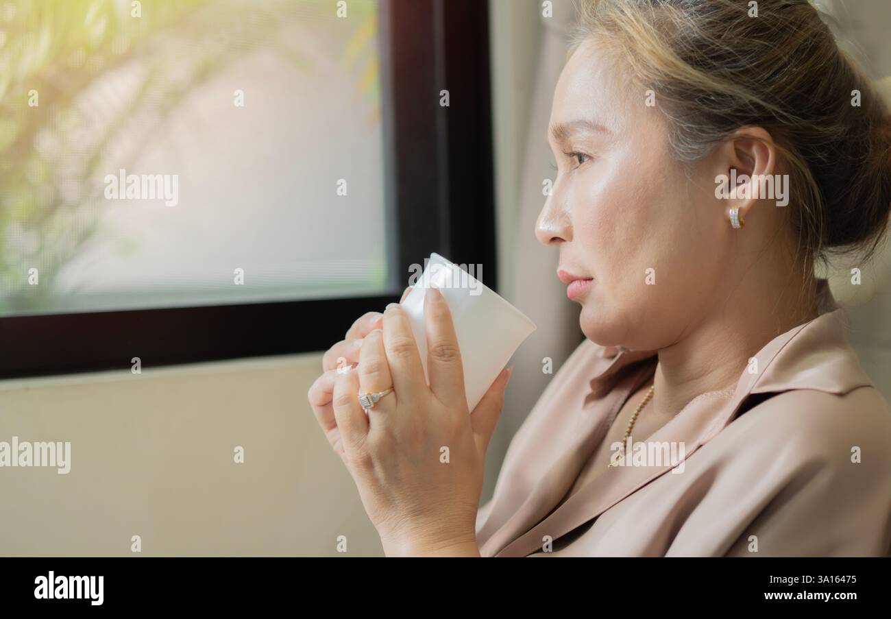 Concerned sad Asian woman with cup of tea sit in living room neayby the ...