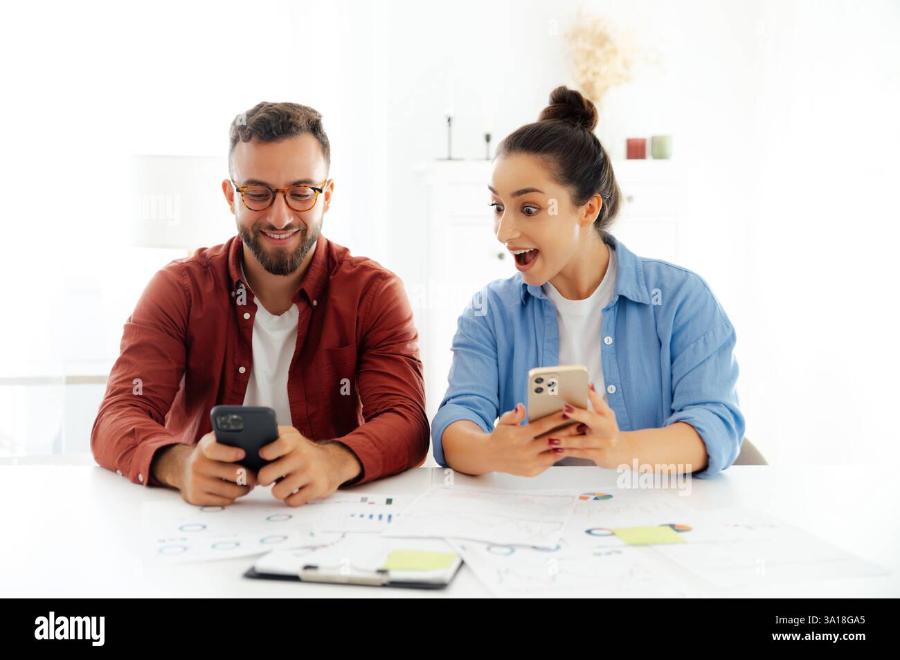Excited man and woman sitting at a desk at office, using their smartphones, reading unexpected news, searching information, demonstrations happy emotions and amazement Stock Photo