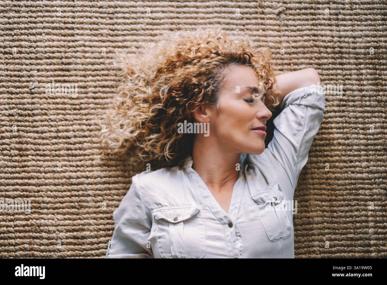 Relaxed and melancholic adult blonde curly woman lying on carpet floor at home looking and thinking. Mindfulness exercise and healthy lifestyle for women enjoying free time meditating Stock Photo