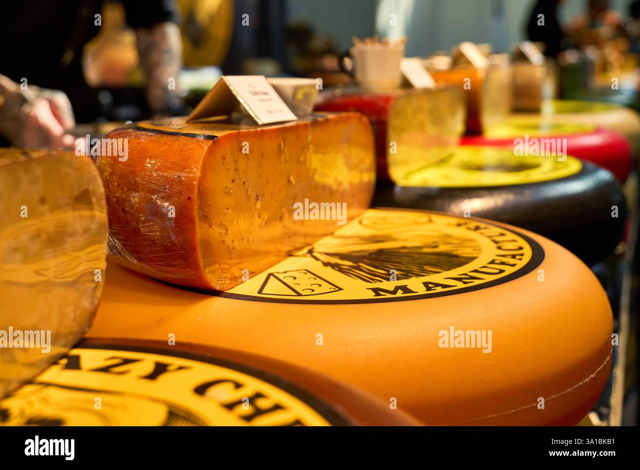 Augsburg, Bavaria, Germany - March 7, 2025: A large cheese wheel with a sliced cheese lies on a sales counter.Other wheels of cheese with labels can be seen in the background *** Ein großes Käserad mit einer aufgeschnittenen Käsesorte liegt auf einer Verkaufstheke. Im Hintergrund sind weitere Käselaibe mit Etiketten zu sehen Stock Photo