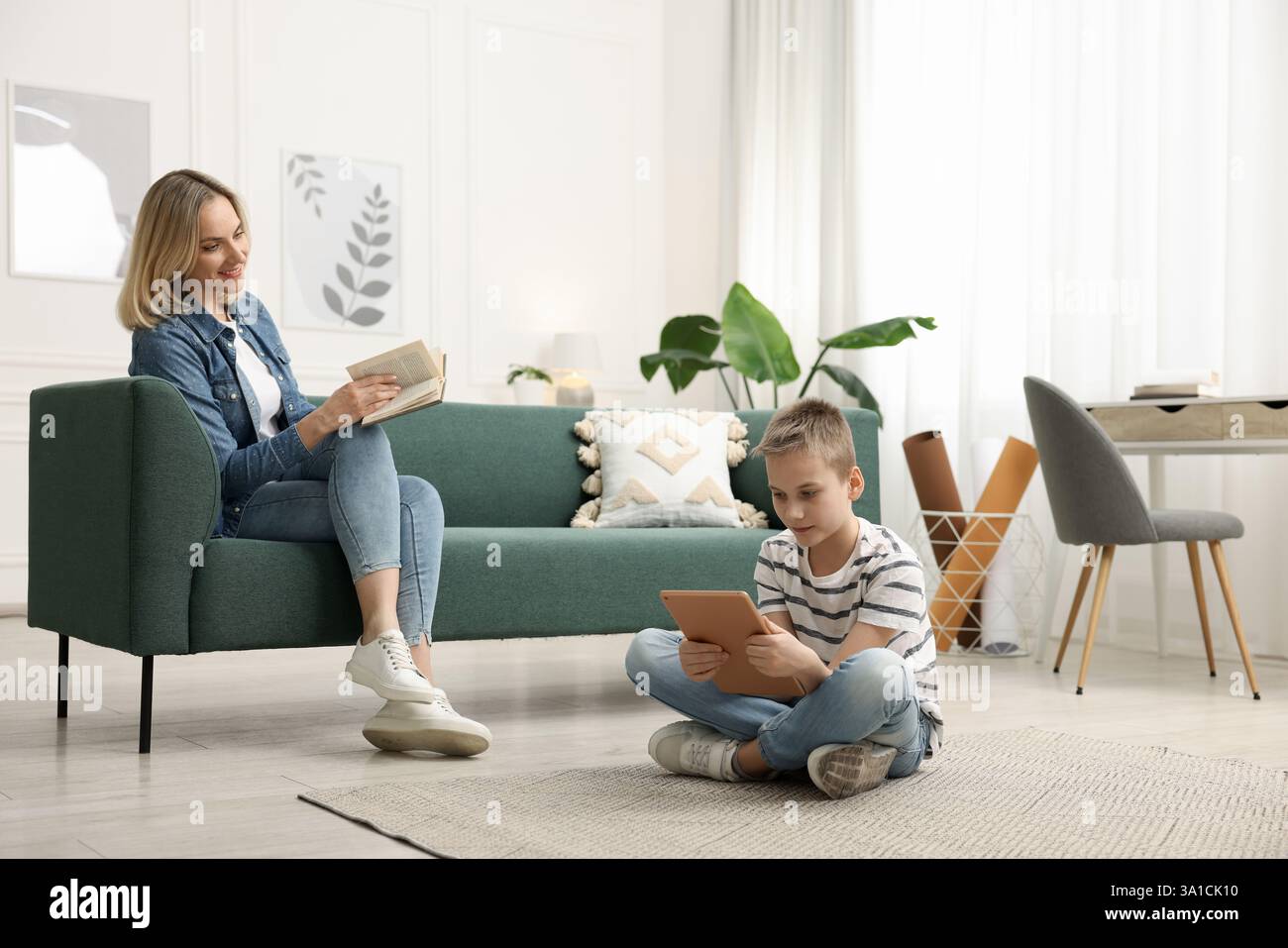 Son using tablet while his mother reading book at home Stock Photo