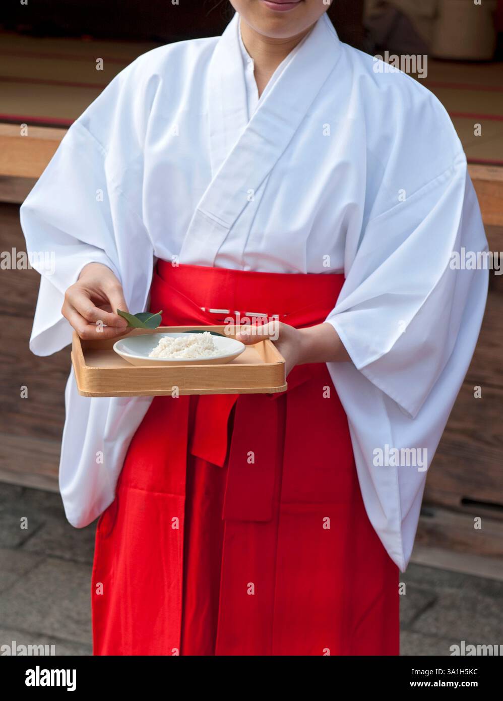 Miko shrine maiden in traditional white kimono and red hakama performing sacred Shinto ritual with sakaki leaves and ceremonial rice offering in Kyoto temple