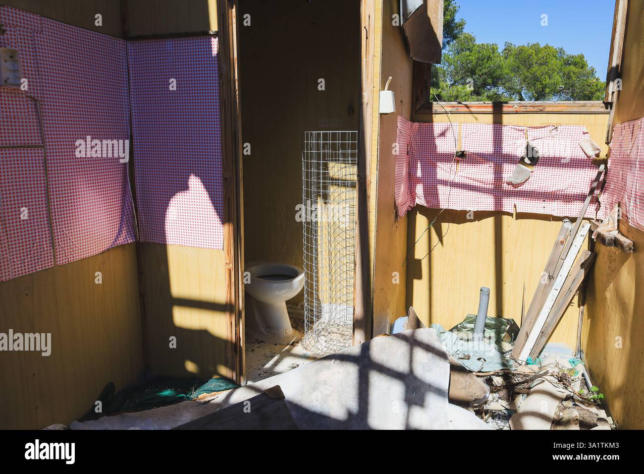 shadows inside a decayed wooden shack, revealing a broken toilet and torn checkered fabric on the walls Stock Photo