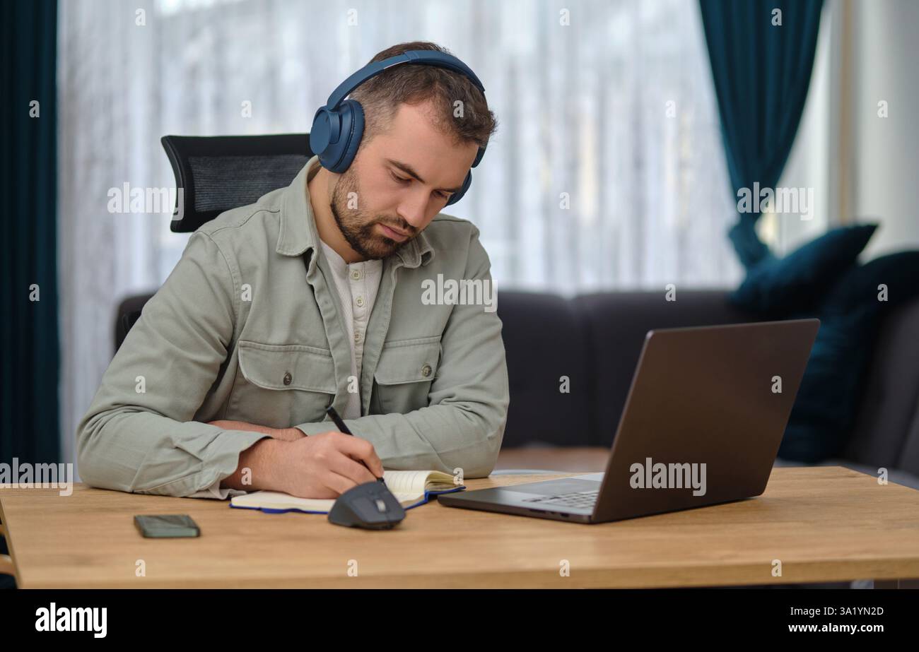 A young man working at home office, wearing wireless headphones, having a video call on a laptop, writing notes, learning webinar or online education. Stock Photo