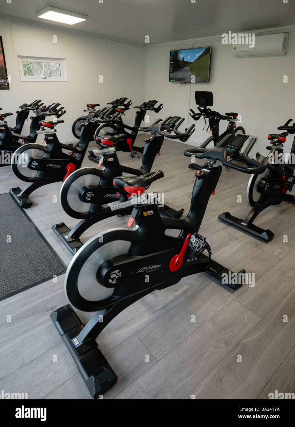 A row of stationary spin bikes are lined up in a gym. The bikes are all the same model and are all in use Stock Photo