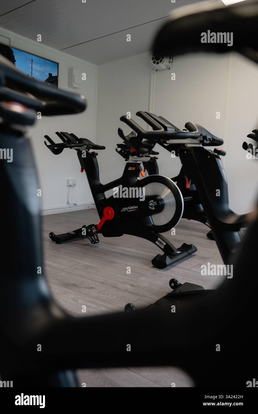 A spin bike is in a gym with a television in the background. The bike is black and has a red and white logo Stock Photo