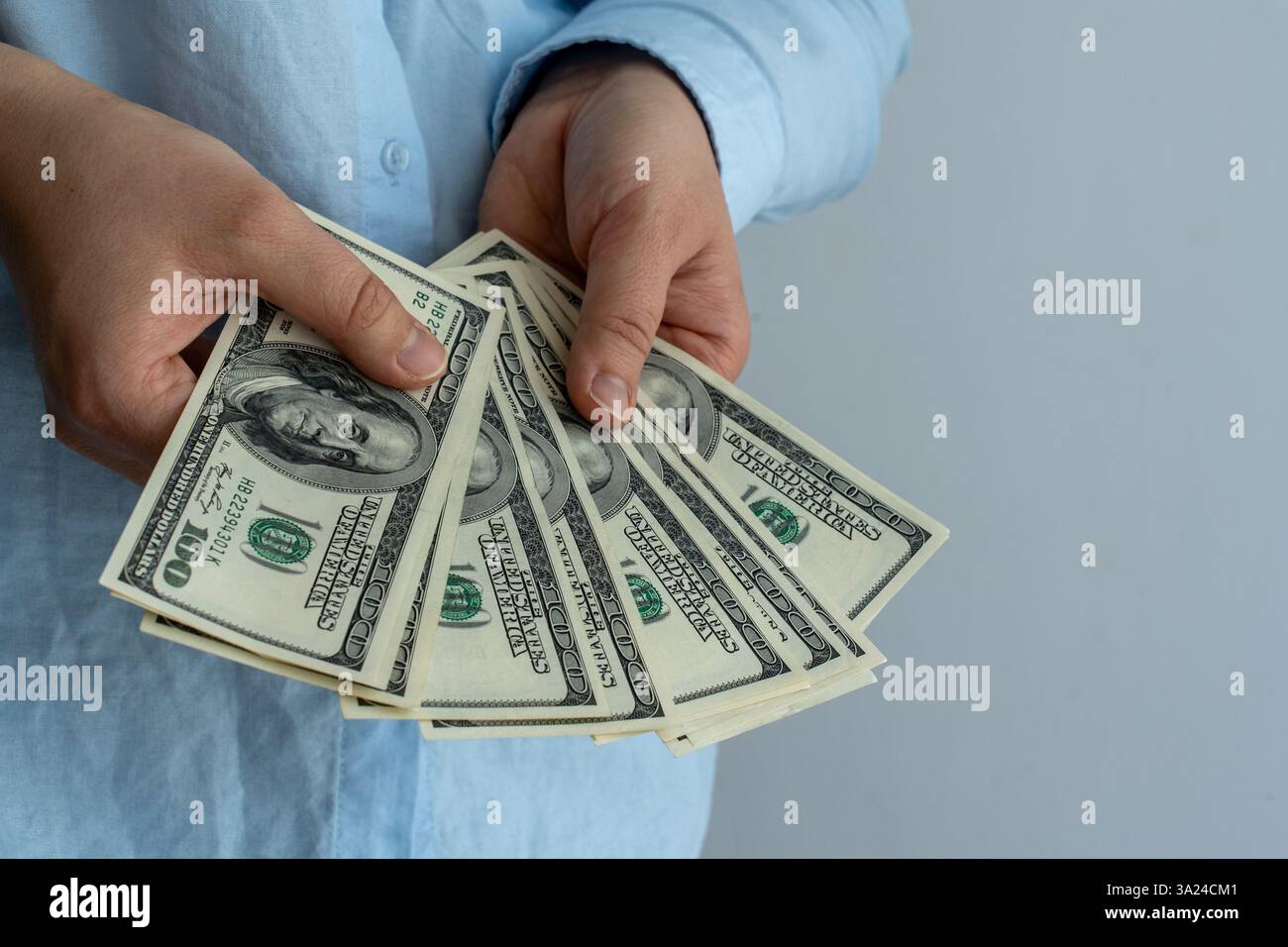 Close-up of a person in a light blue shirt holding multiple US hundred-dollar bills in both hands, fanned out. Concept of financial success, wealth, i Stock Photo