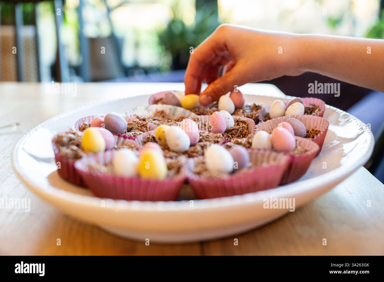Hand putting decorations on to Easter cup cakes in dish on kitchen table. Stock Photo
