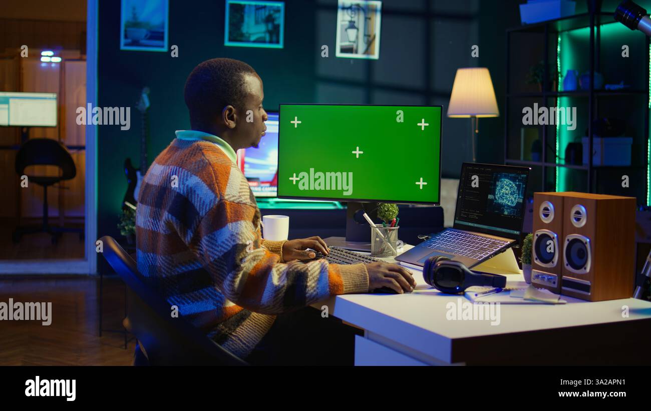 Black person programming neural network code with a green screen on computer to build databases. System engineer working from home while looking at data center security servers. Camera B. Stock Photo