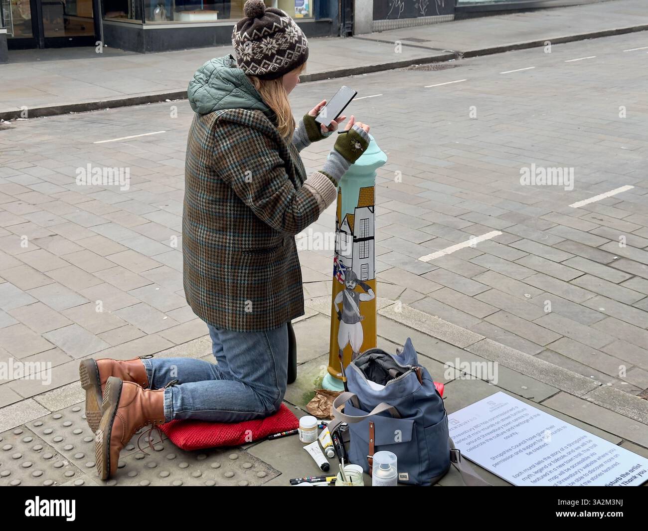 Young female artist, dressed for the cold, kneels beside a kerbside bollard while painting it to decorate it. - Smartphone Captured Stock Image
