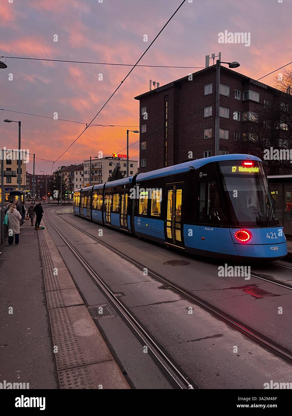 Caption: A blue Oslo tram moves through the city at sunset, its glowing interior lights contrasting with the dramatic pink and orange sky. - Smartphone Captured Stock Image