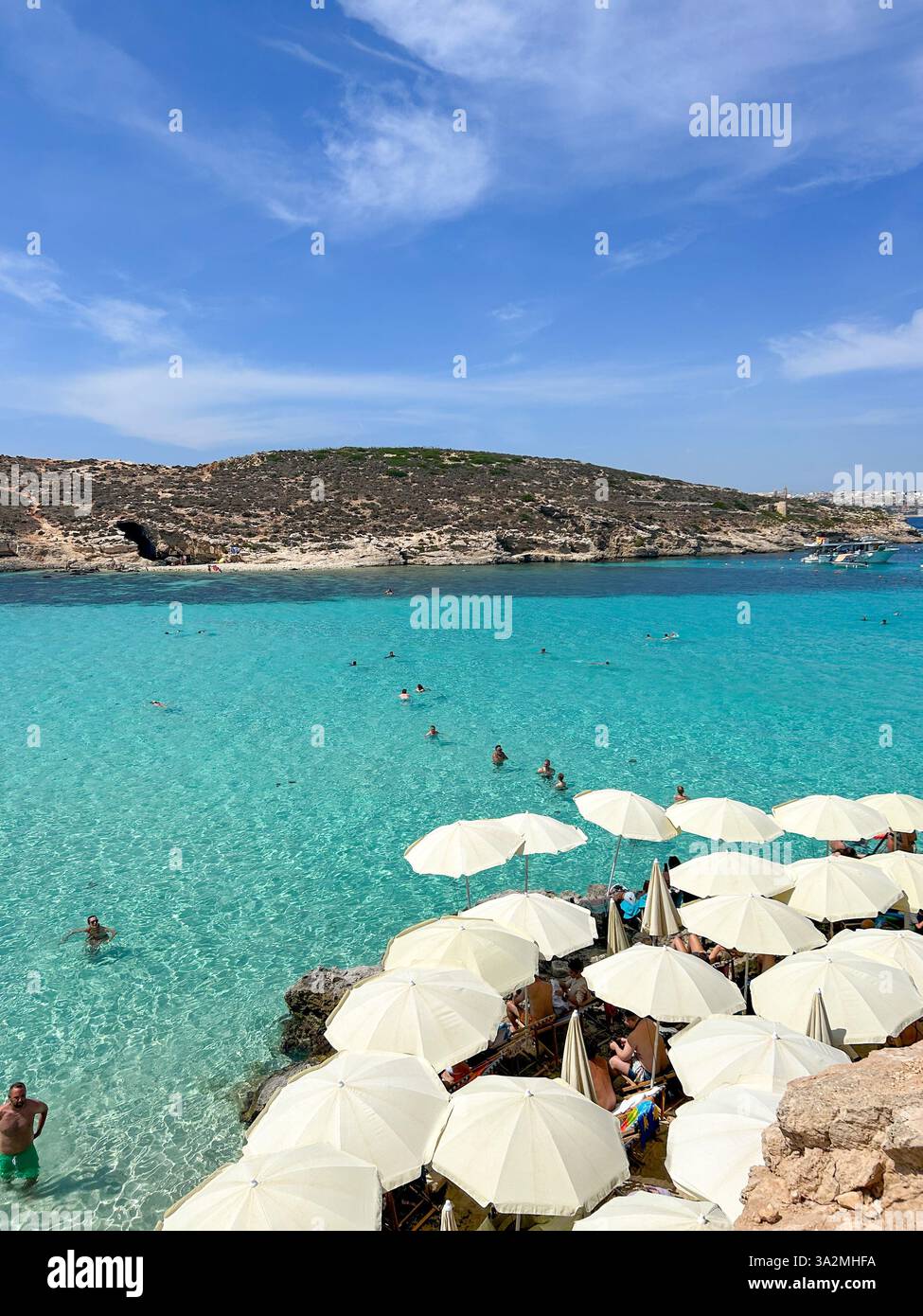 A beautiful beach in the Blue Lagoon, Malta, with white sun umbrellas lining the shore and swimmers enjoying the crystal-clear water. A perfect summer - Smartphone Captured Stock Image