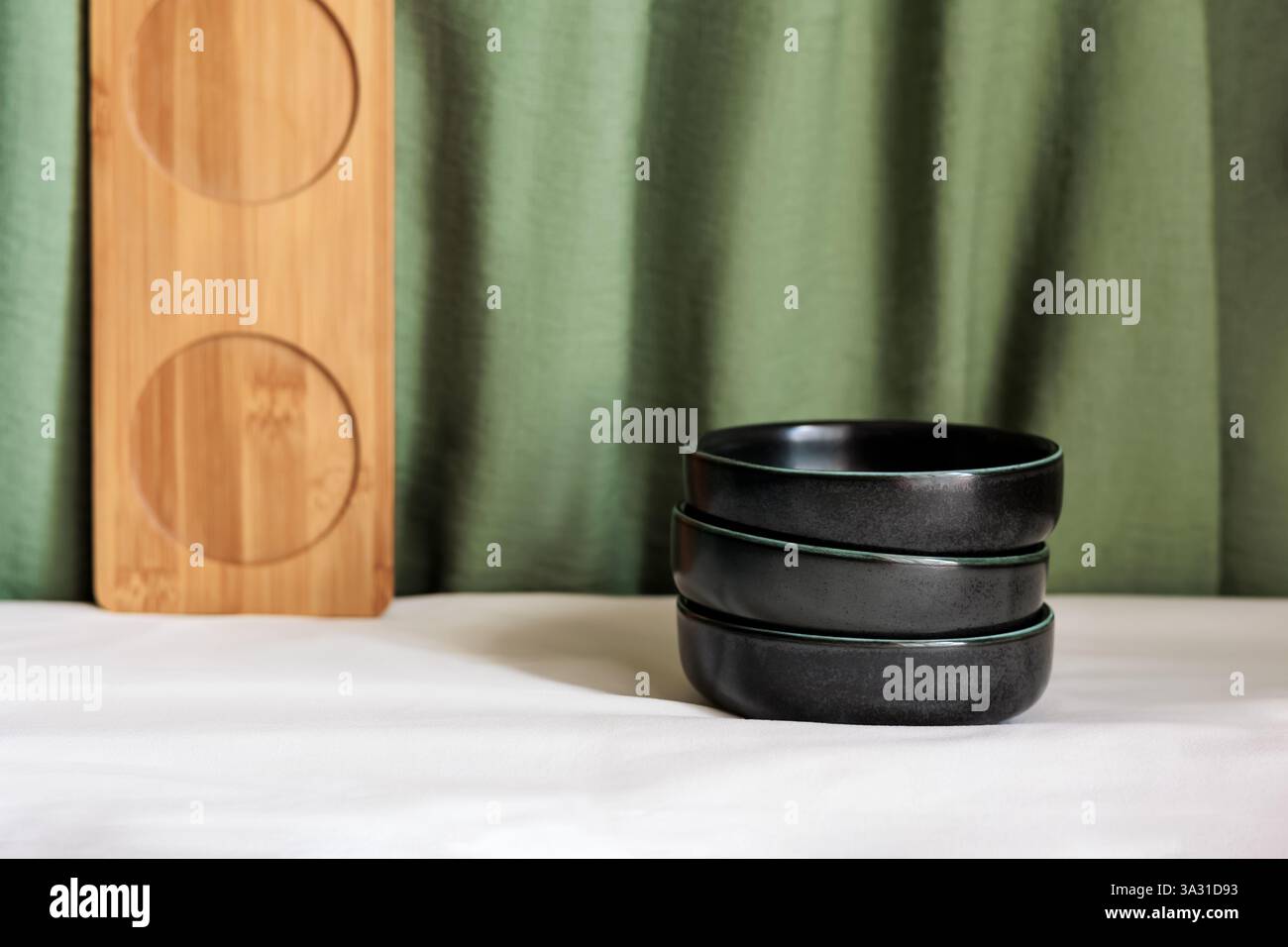 Minimalist composition with stacked black bowls, a wooden tray, and a green fabric backdrop Stock Photo