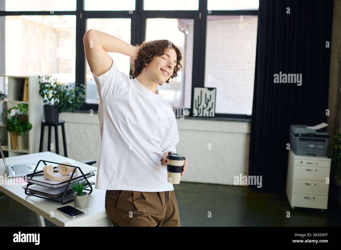 Handsome young man smiles and stretches at his desk during a refreshing coffee break in the office. Stock Photo