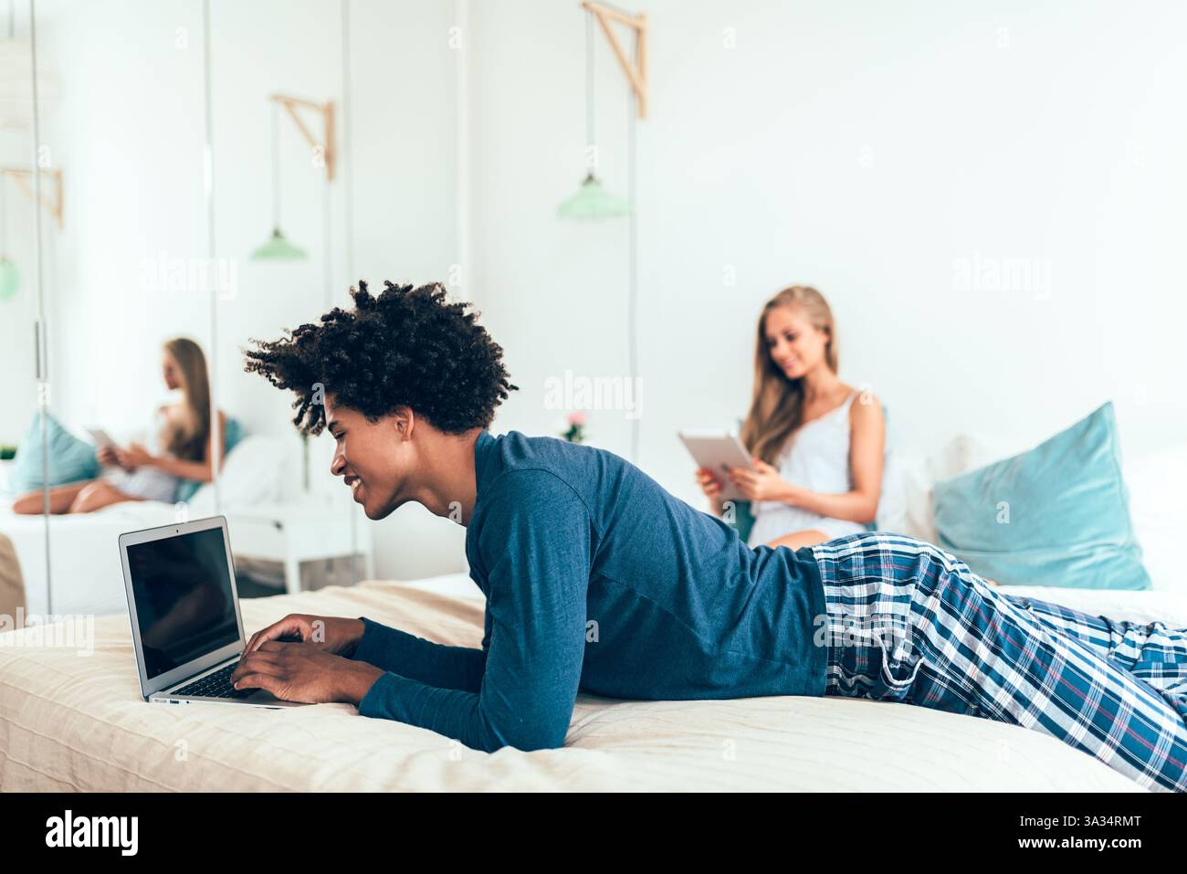 A young man using a laptop and a woman browsing a tablet in a cozy bedroom. They are casually dressed in leisurewear, enjoying a relaxed morning at ho Stock Photo