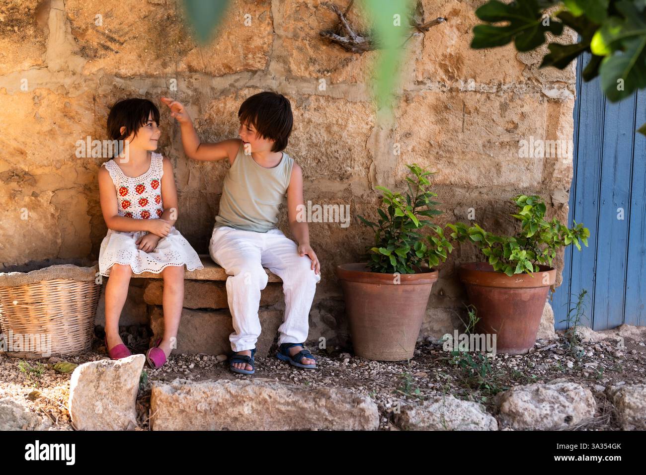 Two children sit on a stone bench near a rustic wall, with potted ...