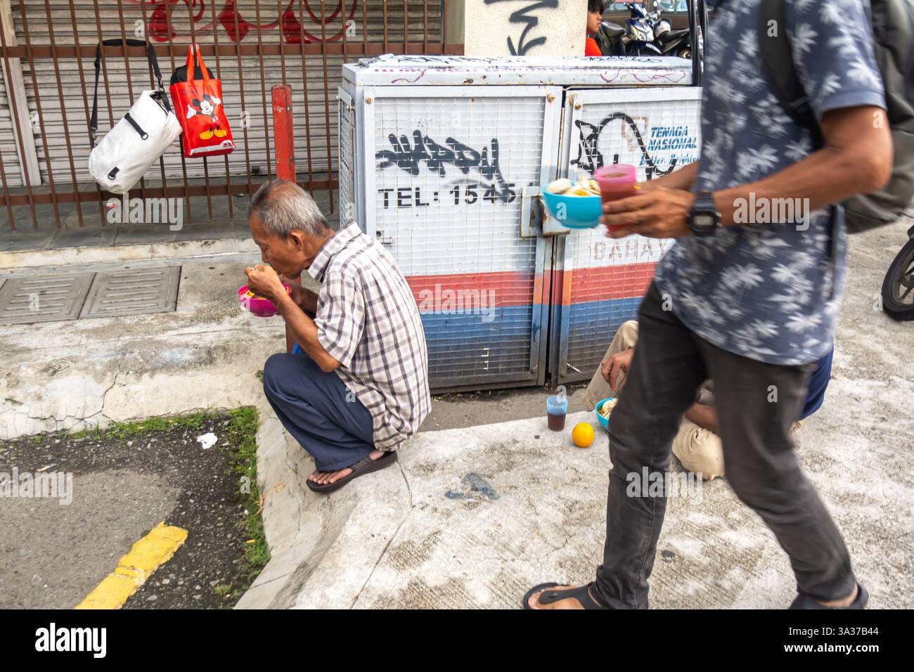 a senior man crouching and holding a food cup eating. A person walking by holding a cup of food, street scene, Chinatown Kuala Lumpur, Malaysia, Asia Stock Photo