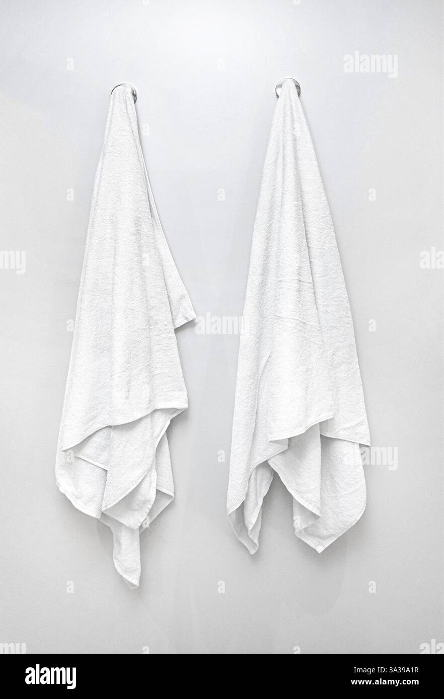Two fresh white towels hanging on hooks in a bathroom, representing ...