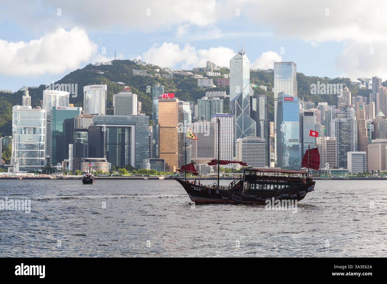 Hong Kong - July 13, 2017: A traditional Chinese boat with red sails cruises through Hong Kong harbor with urban skyscrapers and lush mountains in the Stock Photo