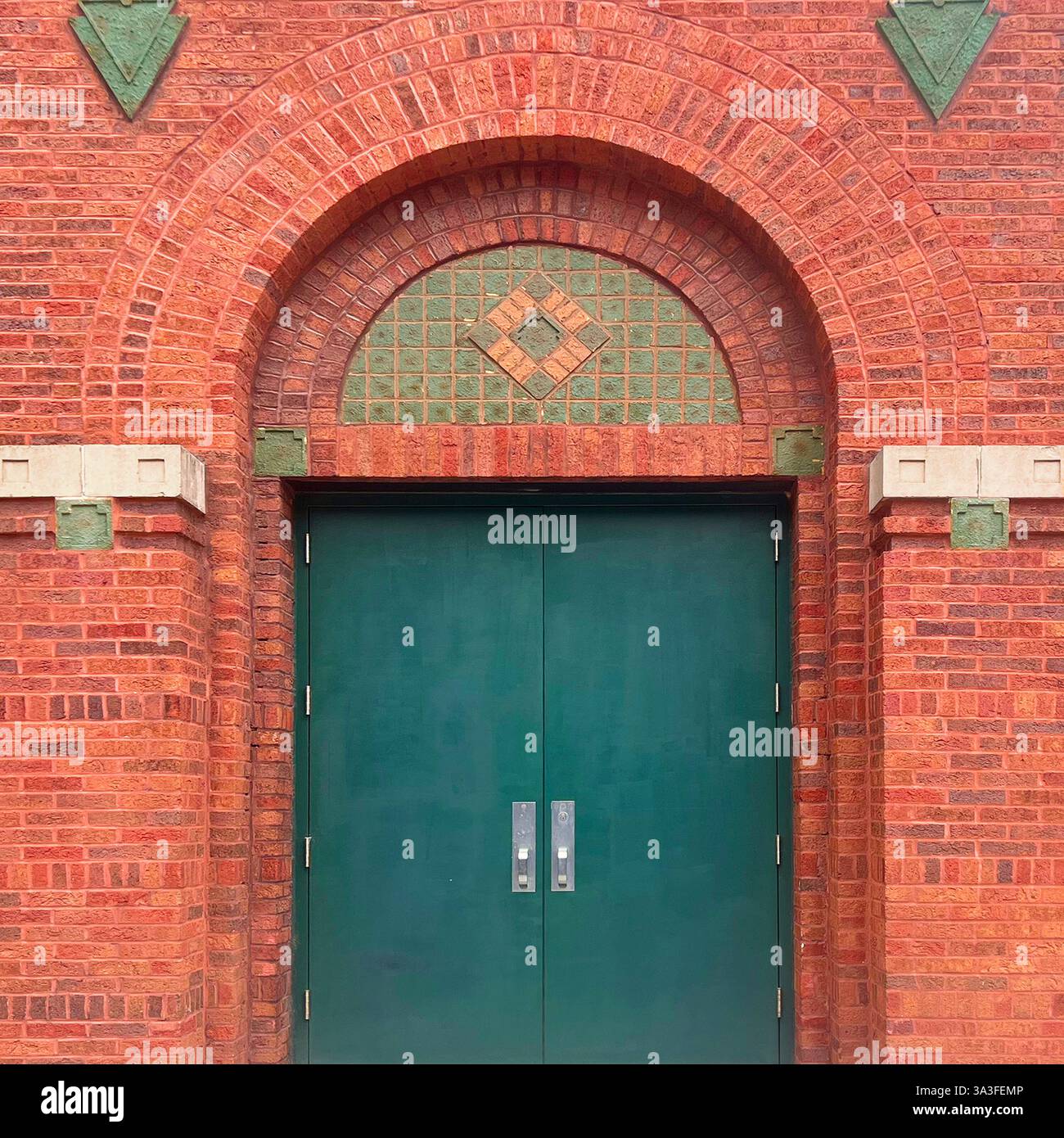 Green double doors set within an arched red brick facade, featuring geometric brick patterns, decorative green tile inlays. - Smartphone Captured Stock Image