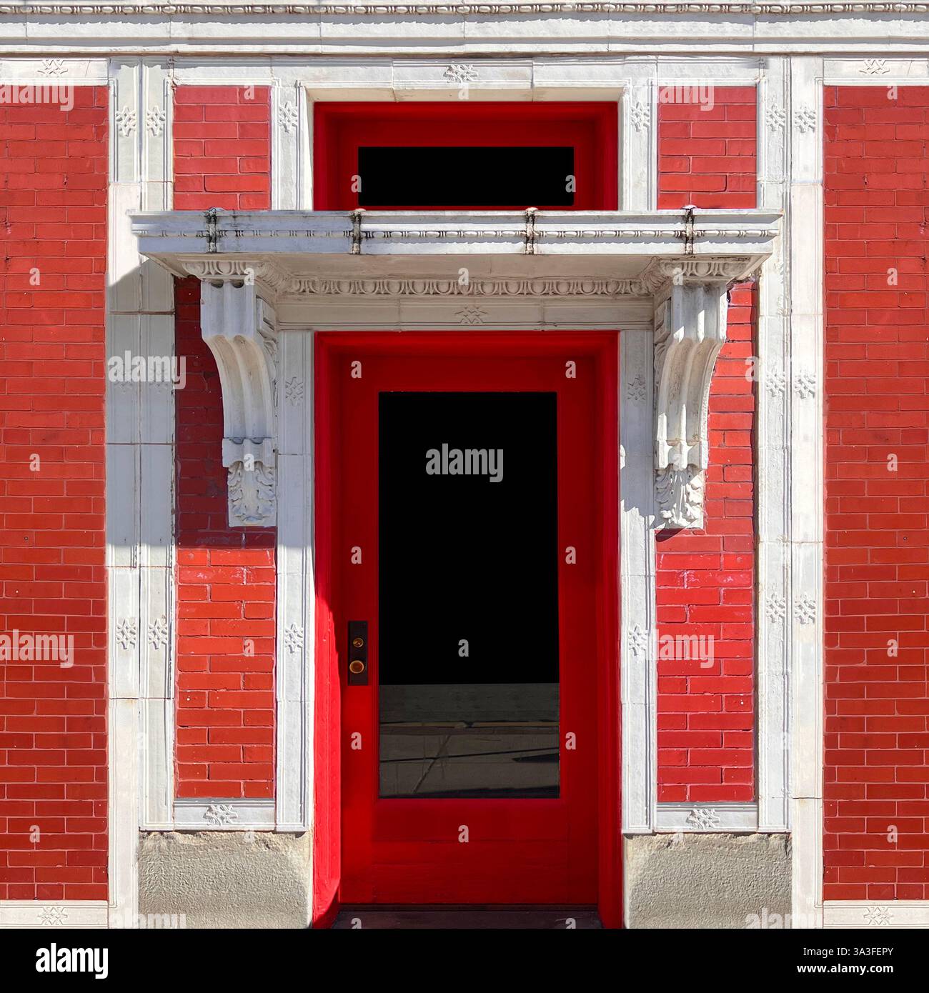 Red door is set within a red brick facade, framed by elegant white stone detailing and an intricately carved overhang and decorative corbels, - Smartphone Captured Stock Image