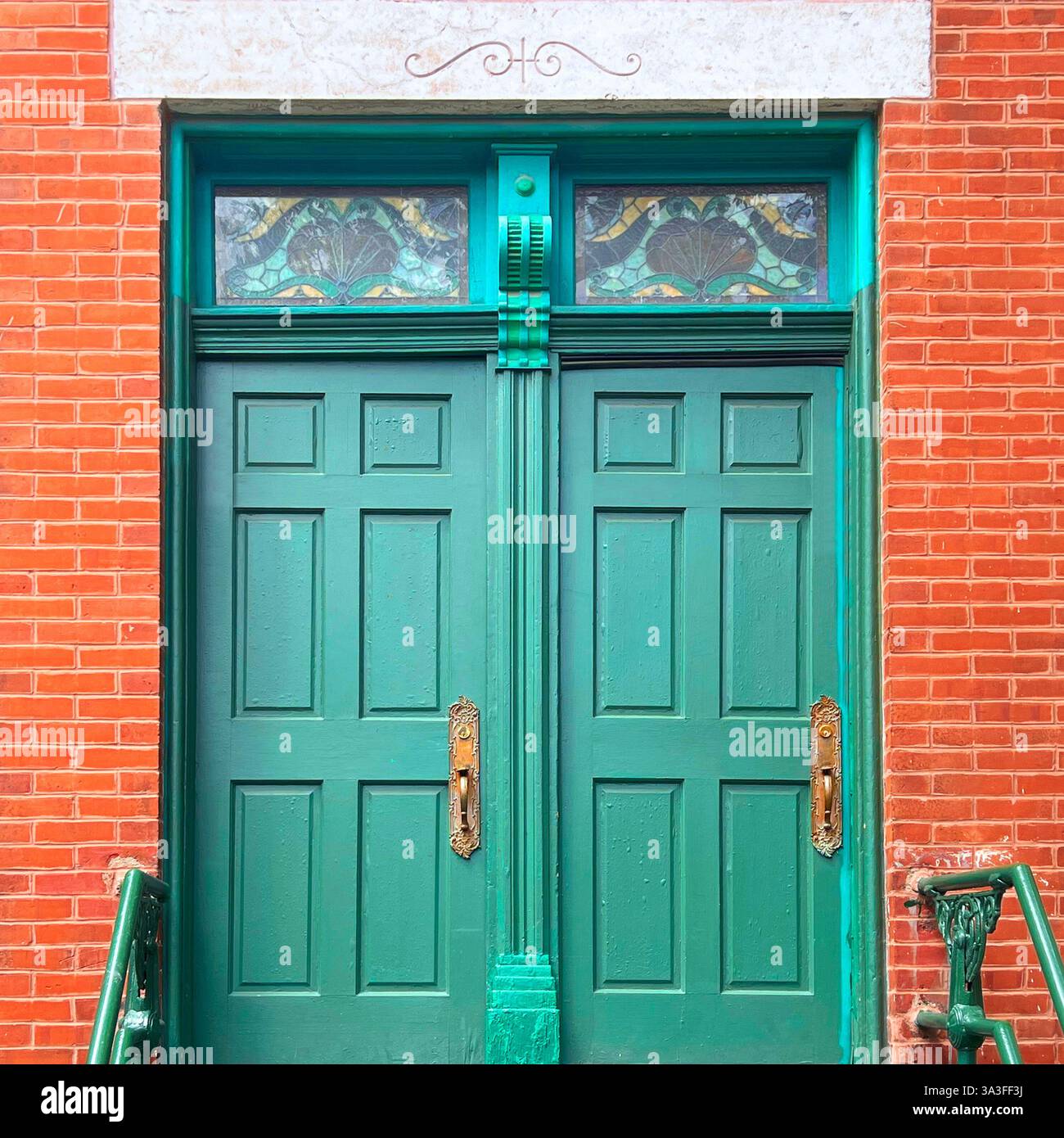 Elegant double green doors with intricate brass handles and stained glass transom windows, set in a historic orange brick building. - Smartphone Captured Stock Image