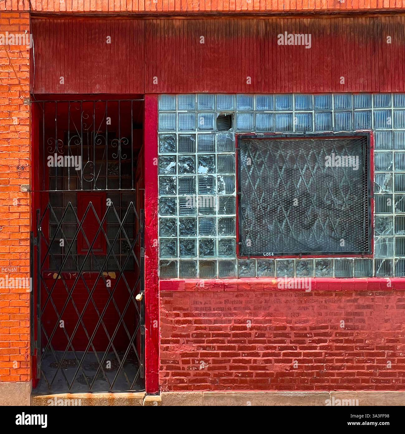 A gated red entrance with a glass block window, covered by a metal security grille, set in a weathered red brick building in an urban environment. - Smartphone Captured Stock Image