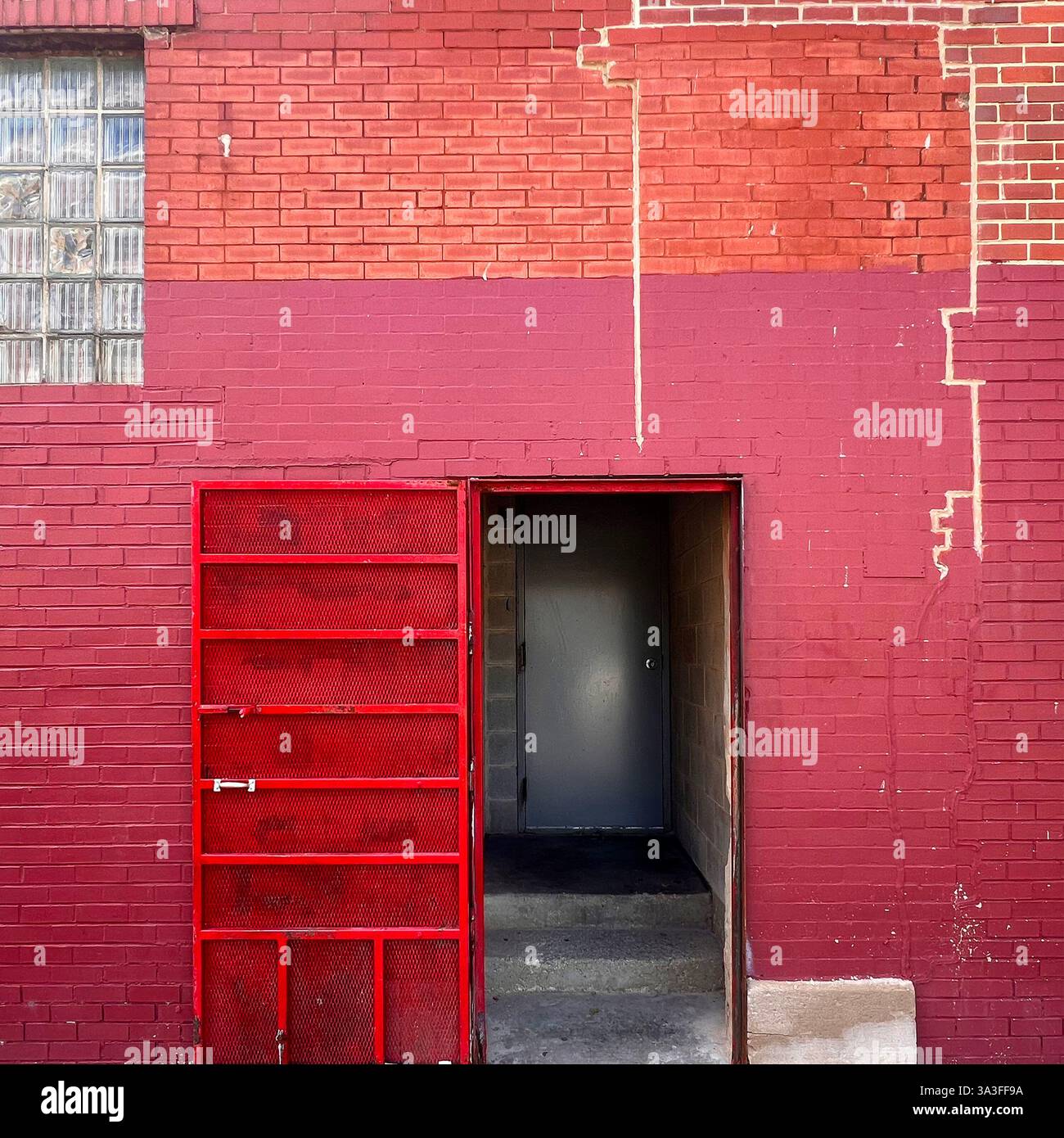 A red metal security gate revealing a dark industrial entrance, set against a weathered red brick wall with visible cracks. - Smartphone Captured Stock Image