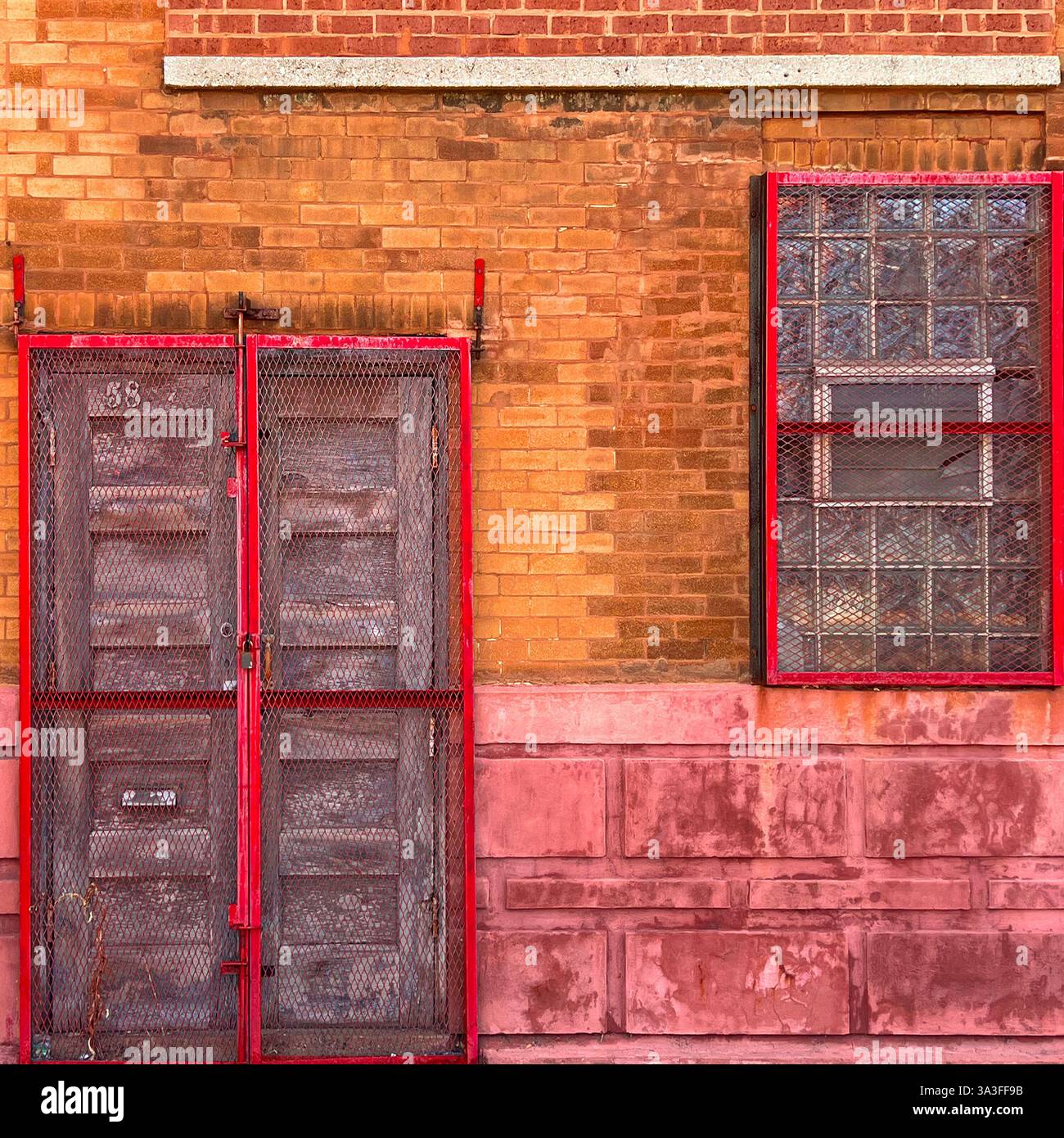 A rustic wooden double door with red metal security mesh, set against a weathered brick wall with a matching window. - Smartphone Captured Stock Image