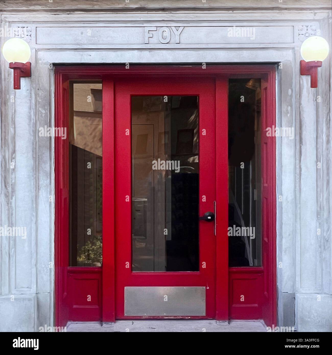 A striking red entrance door set within a historic stone facade, featuring the engraved name 'FOY' above, flanked by two vintage globe lights - Smartphone Captured Stock Image