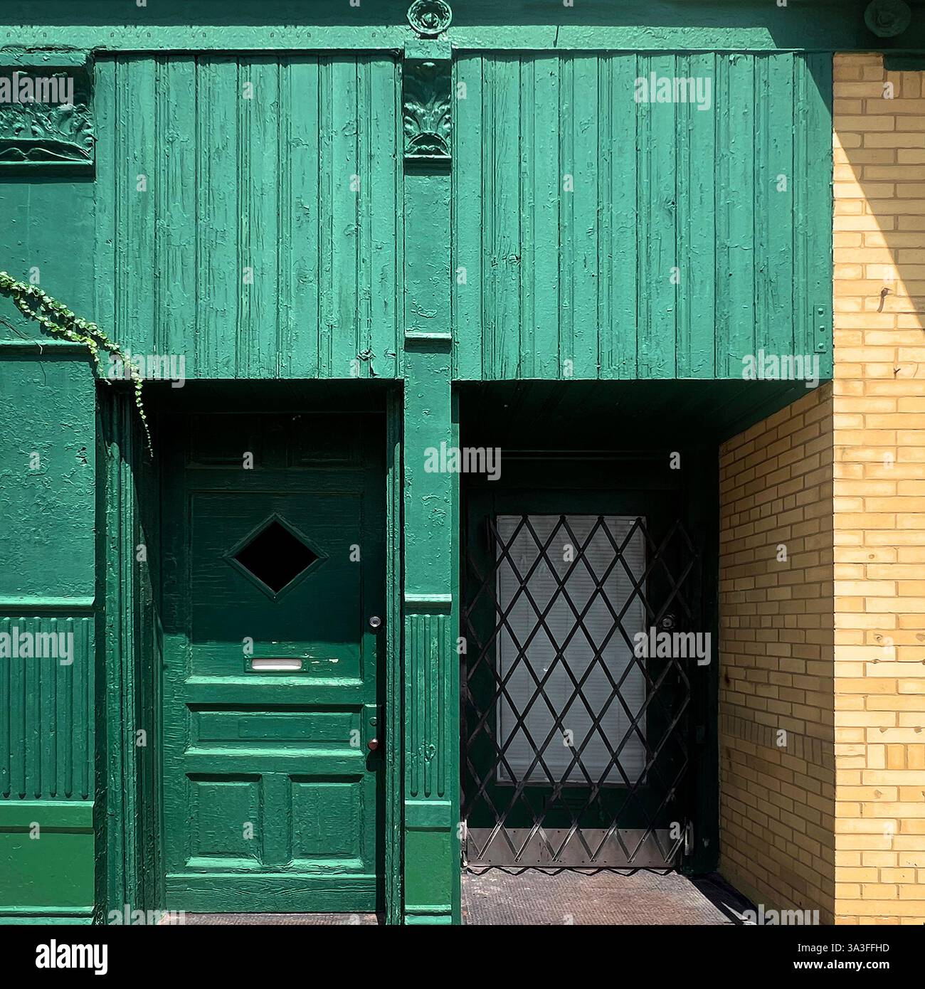 Green wooden storefront with a vintage door featuring a diamond-shaped window and an adjacent gated entrance, bathed in sunlight against yellow brick - Smartphone Captured Stock Image