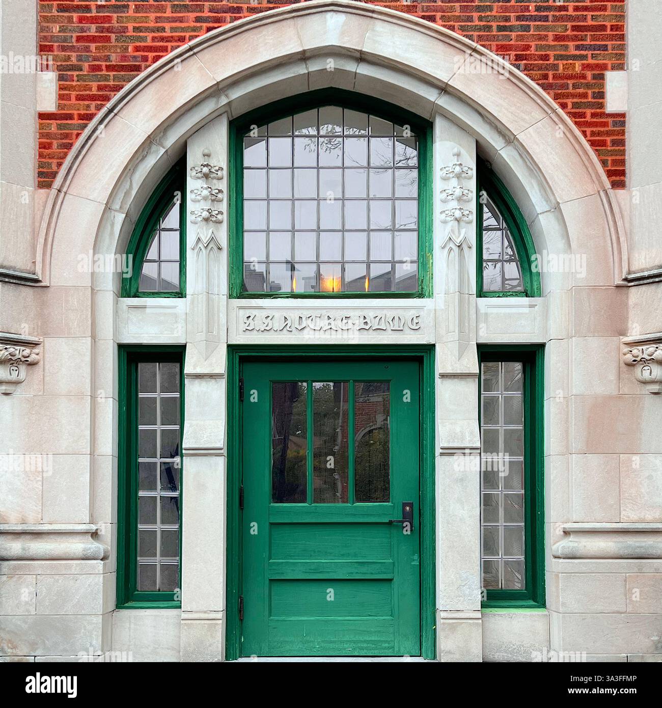 Gothic-style green door with an arched limestone entryway, intricate stone detailing, and large glass windows, set against a red brick facade. - Smartphone Captured Stock Image