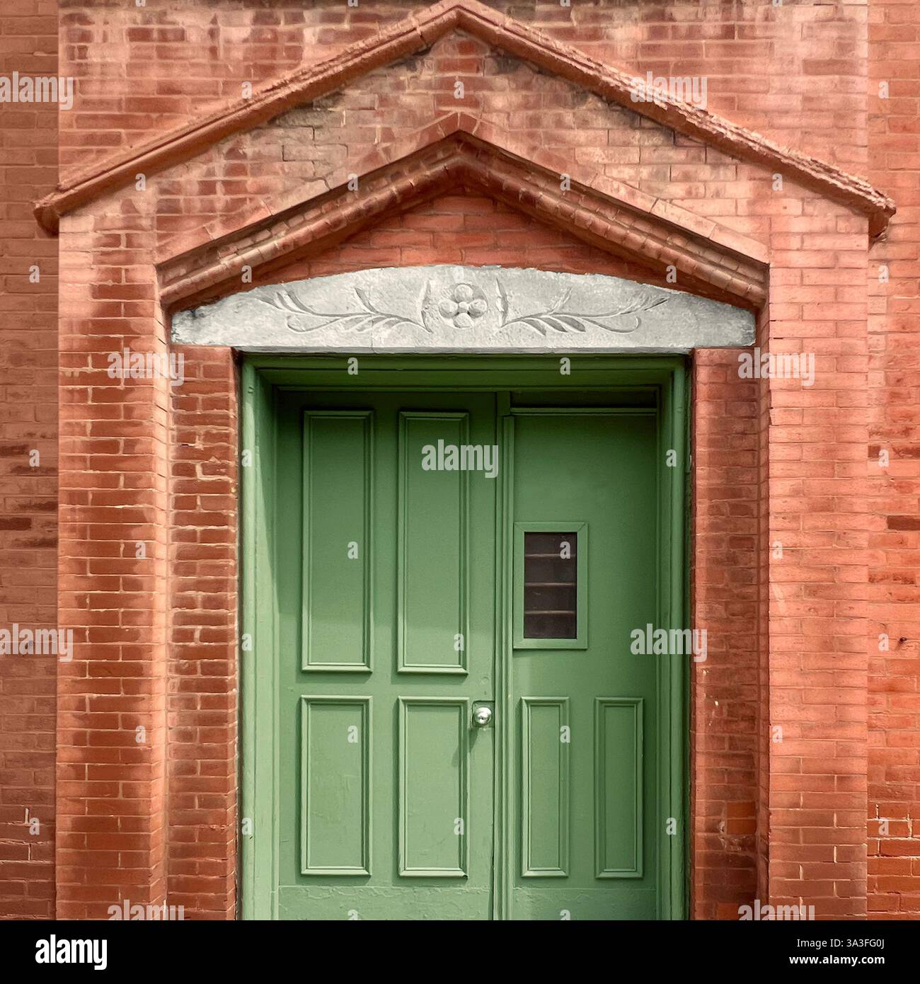 Green door set within a red brick facade, framed by an arched brick structure and an ornate stone lintel with floral carvings - Smartphone Captured Stock Image