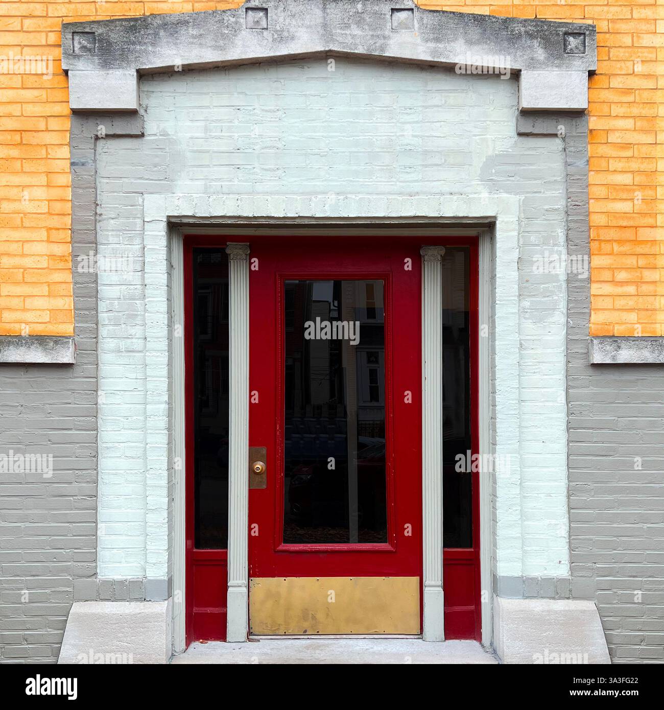 A red door with decorative columns and a brass kick plate, framed by a muted brick facade with yellow and gray tones, showcasing vintage architectural - Smartphone Captured Stock Image