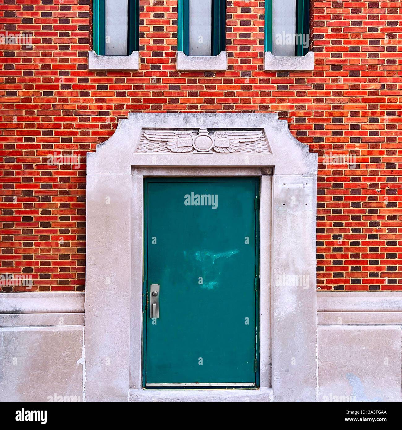 Art Deco Teal green door set in decorative stone doorway with carved Egyptian Revival motifs against vibrant red brick wall, Chicago. - Smartphone Captured Stock Image