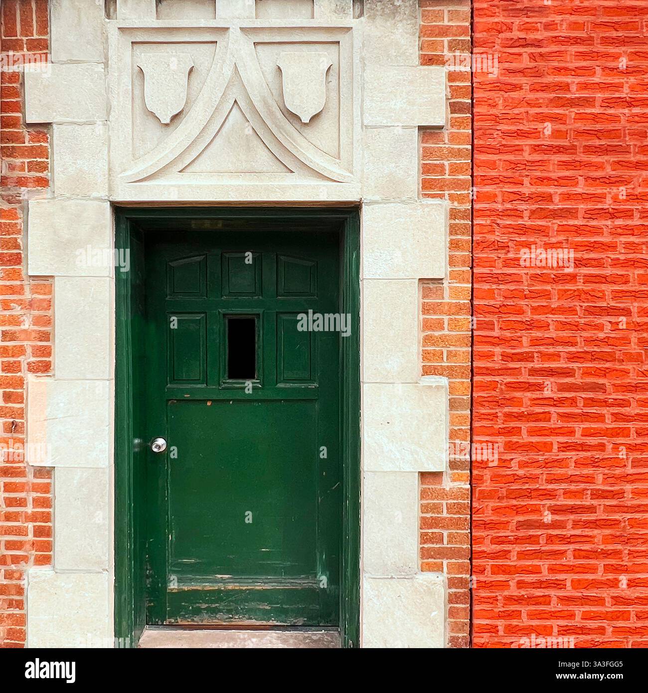 Green door with a small window, set within an ornate stone frame featuring decorative shield motifs, surrounded by ornage brick facade - Smartphone Captured Stock Image