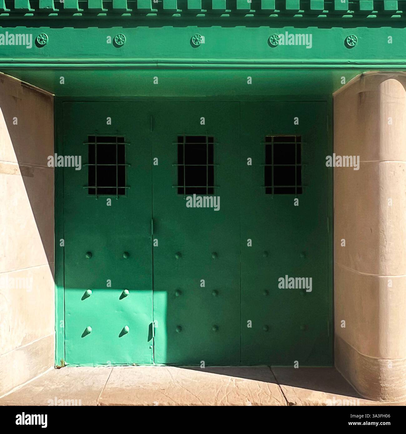 A green industrial-style metal door with rivets and small barred windows, framed by stone columns and partially illuminated by sunlight in Chicago - Smartphone Captured Stock Image