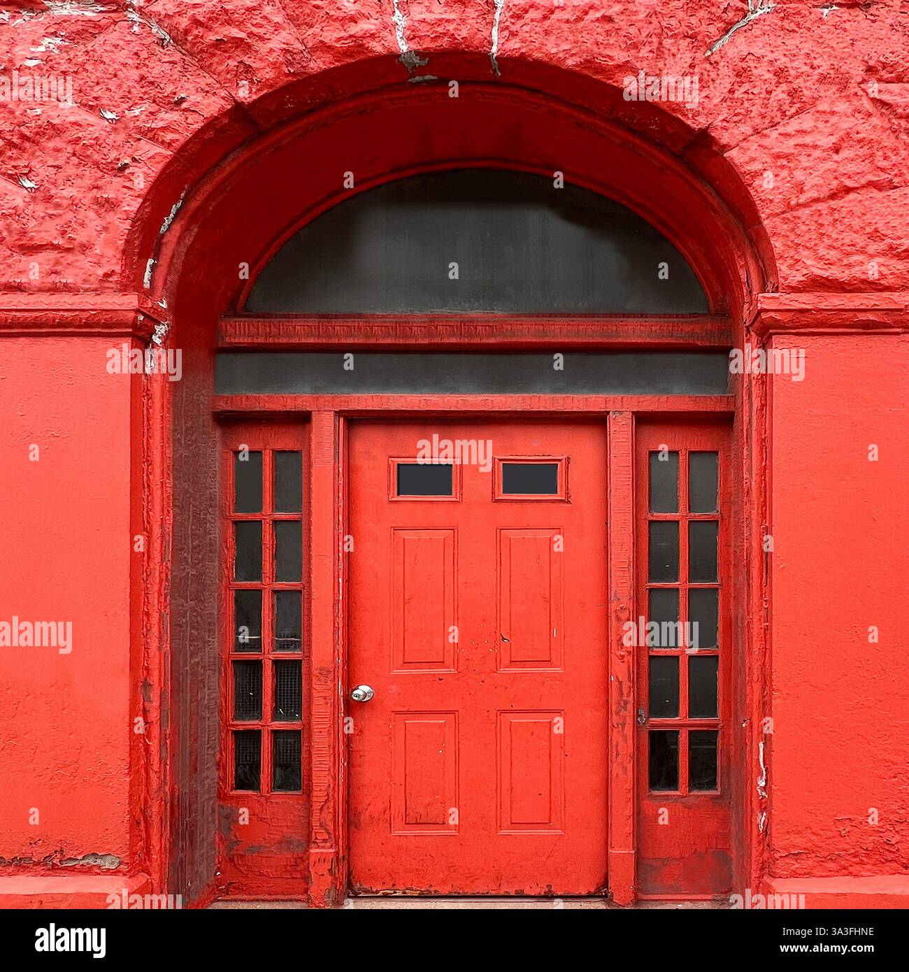 Red door with transom and sidelights set within a textured red arched facade, exuding a weathered yet striking urban charm. - Smartphone Captured Stock Image
