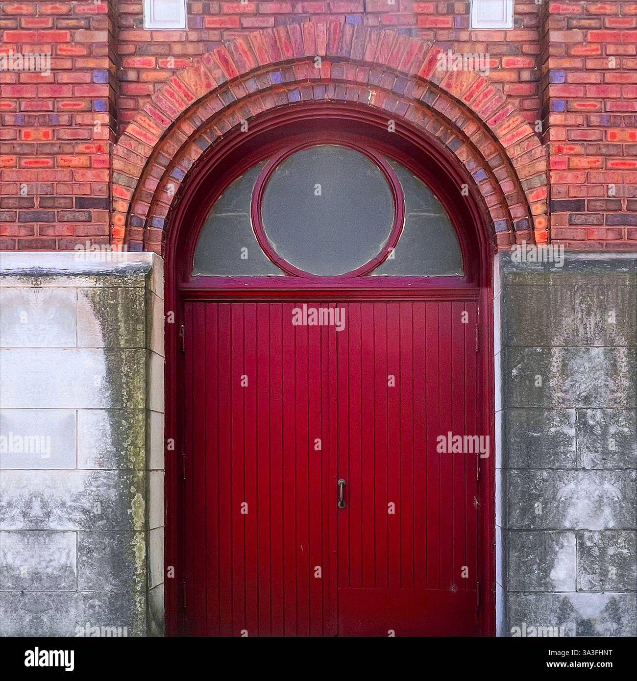 A historic red door with an arched transom window, set within a brick and stone facade,entrance features semi-circular glass panel - Smartphone Captured Stock Image