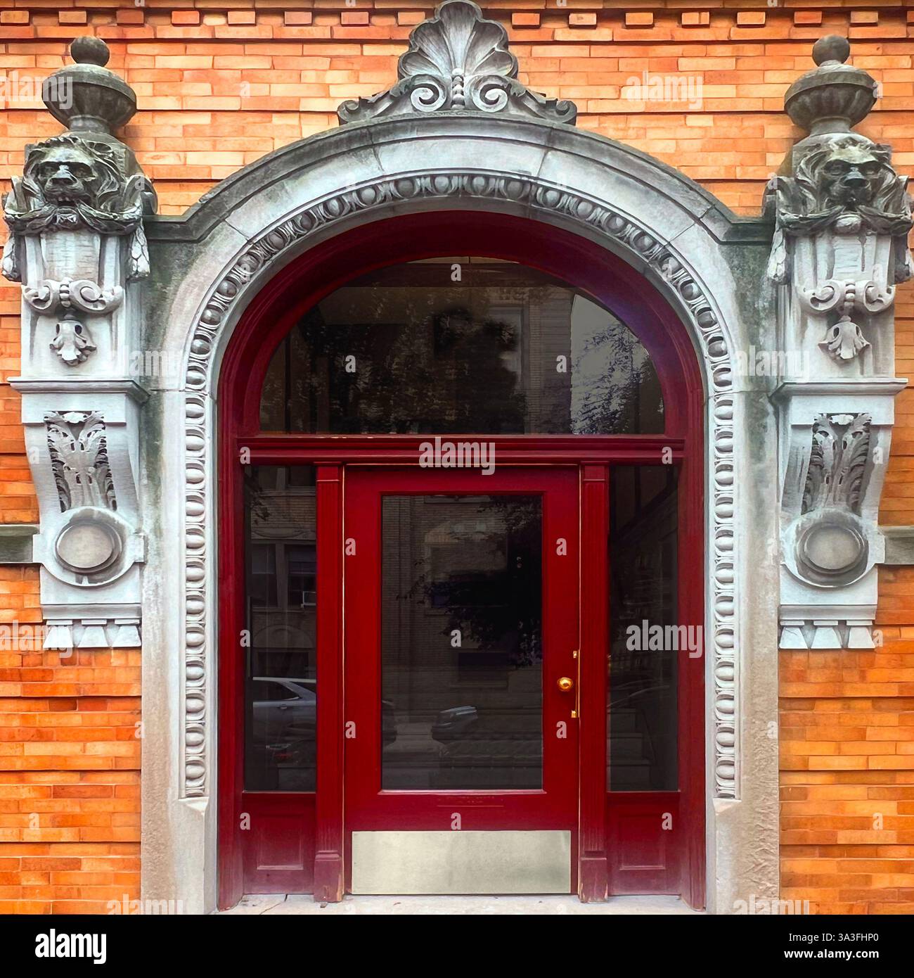 Elegant red door framed by an intricate stone archway adorned with sculpted lion heads and decorative elements, set against a warm brick facade - Smartphone Captured Stock Image