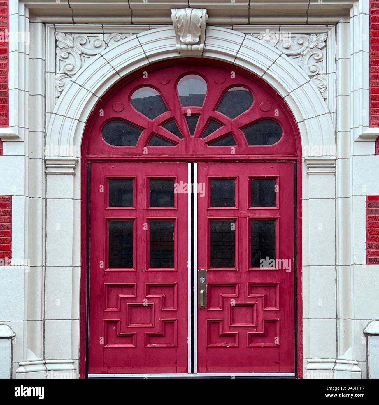 Elegant red double doors with a fanlight window and intricate geometric paneling, framed by a decorative white stone archway on a historic red brick. - Smartphone Captured Stock Image